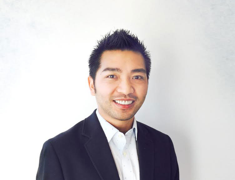 A man with a broad smile is wearing a dark suit and standing in front of a white background.